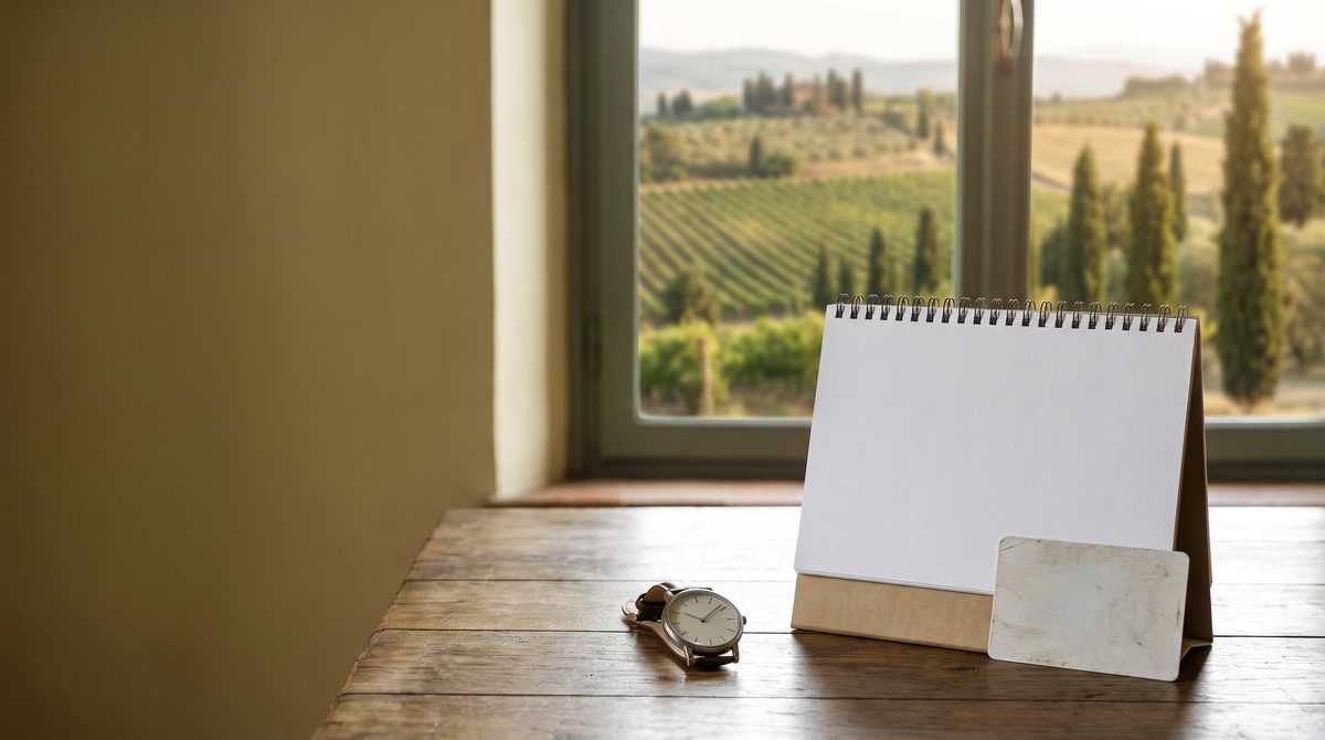 Un tavolo di legno con un calendario da tavolo vuoto, un orologio e una carta bianca, con vista su un paesaggio di vigneti.