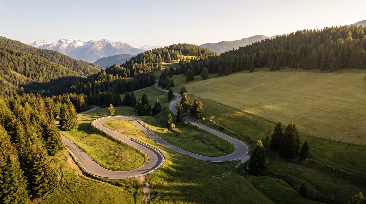 Una strada di montagna serpeggiante attraversa una foresta lussureggiante con cime innevate sullo sfondo sotto un cielo limpido.
